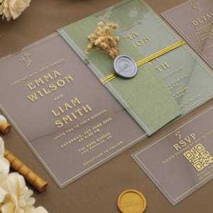 May include: Elegant wedding invitation suite featuring clear acrylic cards with gold lettering. The suite includes details, RSVP, and invitation cards with names and wedding details. A green card is adorned with dried flowers and a wax seal.