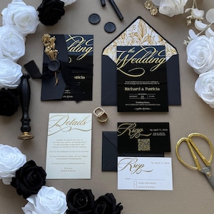 May include: A black and gold wedding invitation suite featuring a main card with gold foil text, an RSVP card with a QR code, a details card, and black envelopes with gold floral liners. Includes a wax seal, a stamp, and gold-toned scissors.