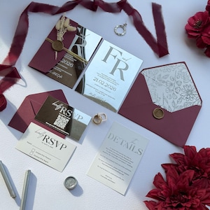 Può includere: Inviti di nozze bordeaux con dettagli a specchio argentati, con le iniziali "FR" e la data "21.02.2026". Il set include biglietti RSVP, buste ed elementi decorativi come fiori secchi e sigilli di cera. Sono presenti anche un anello e un nastro bordeaux.