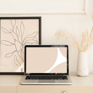 Puede incluir: Un ordenador portátil con un diseño abstracto beige y rosa en la pantalla está sentado sobre una superficie marrón claro. Un cuadro negro enmarcado con un dibujo lineal de una planta está detrás del portátil. Un jarrón con flores secas está a la derecha del portátil.