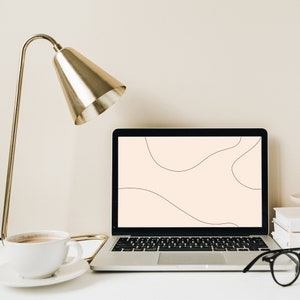 Pode incluir: Uma lâmpada de mesa dourada com um computador portátil branco em uma mesa branca. A tela do laptop exibe um fundo bege claro com linhas abstratas pretas. Uma caneca de café branca e um par de óculos pretos também estão na mesa.