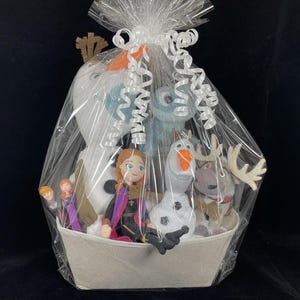 May include: A gift basket filled with Frozen-themed plush toys and dolls. The basket includes a large Olaf snowman, Anna and Elsa dolls, and other characters. The basket is wrapped in clear cellophane and tied with a white ribbon.