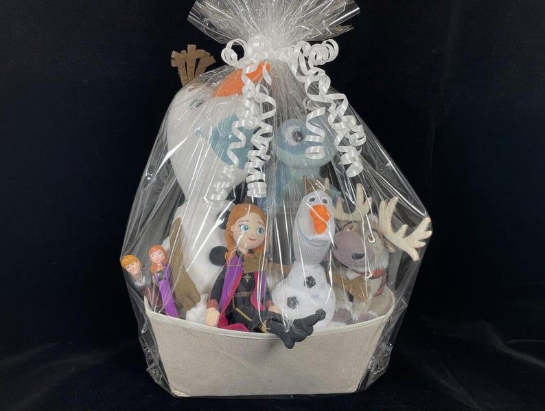 May include: A gift basket filled with Frozen-themed plush toys and dolls. The basket includes a large Olaf snowman, Anna and Elsa dolls, and other characters. The basket is wrapped in clear cellophane and tied with a white ribbon.