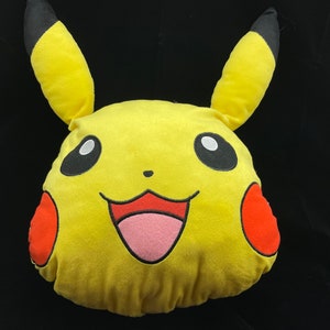 May include: A yellow Pikachu plush pillow with black-tipped ears, red cheek circles, and a smiling mouth. The pillow has a pink tongue and black eyes. The plush is a character from the popular Pokemon franchise.
