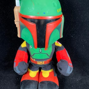 May include: A plush Boba Fett figure, featuring a green helmet with red accents and a black visor. The figure has a green and red torso, red arms, and black feet with red and yellow details. The figure is approximately 25 cm tall.