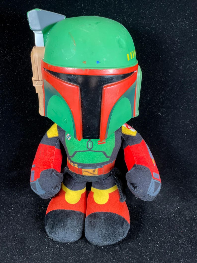 May include: A plush Boba Fett figure, featuring a green helmet with red accents and a black visor. The figure has a green and red torso, red arms, and black feet with red and yellow details. The figure is approximately 25 cm tall.