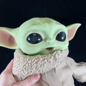 May include: A plush toy of the character Baby Yoda from the Star Wars series The Mandalorian. The toy is green with large eyes and is wearing a brown coat with a white fur collar.