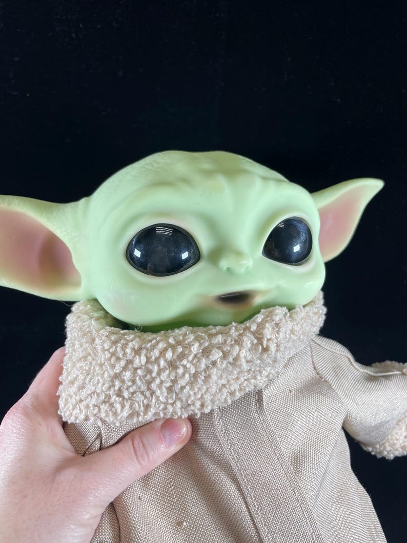 May include: A plush toy of the character Baby Yoda from the Star Wars series The Mandalorian. The toy is green with large eyes and is wearing a brown coat with a white fur collar.