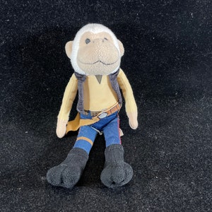 May include: A plush toy monkey dressed as a character from Star Wars. The monkey is wearing a brown vest, a blue belt, and blue jeans. The monkey is sitting with its legs crossed.