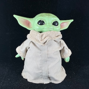 May include: A green plush toy with large ears and big eyes, wearing a brown hooded coat. The toy is a popular character from the Star Wars franchise.