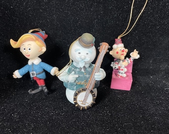 Puede incluir: Tres adornos navideños: un niño con abrigo azul, un muñeco de nieve tocando el banjo y un payaso en una silla rosa. El niño tiene un sombrero amarillo y rojo. El muñeco de nieve lleva un sombrero y un chaleco verde estampado. El payaso viste un traje rojo y blanco.