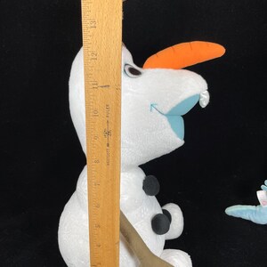 May include: A plush Olaf doll from the movie Frozen, with a carrot nose, black button eyes, and three black buttons on the body. The doll is approximately 10 inches tall, as measured by a wooden ruler.