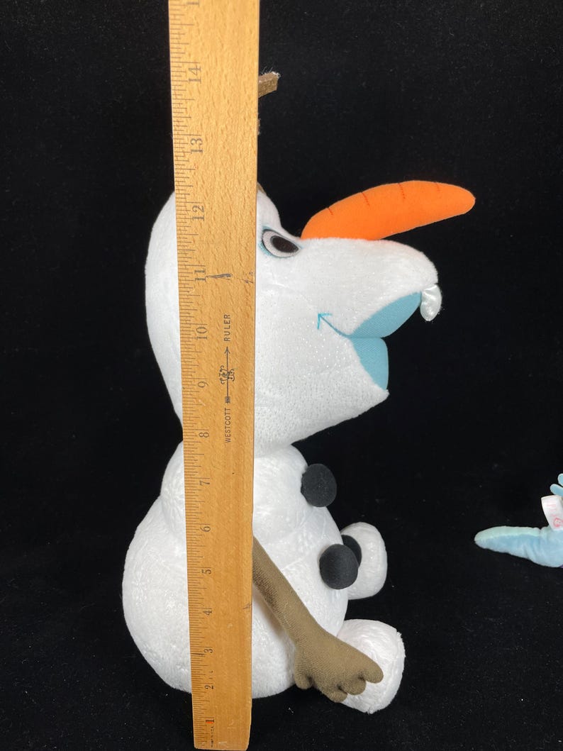 May include: A plush Olaf doll from the movie Frozen, with a carrot nose, black button eyes, and three black buttons on the body. The doll is approximately 10 inches tall, as measured by a wooden ruler.