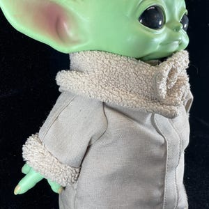May include: A green-skinned doll with large black eyes and pointed ears. The doll is dressed in a tan coat with a fluffy, cream-coloured collar and cuffs. The doll's hand is extended, and the overall appearance is reminiscent of a popular character.