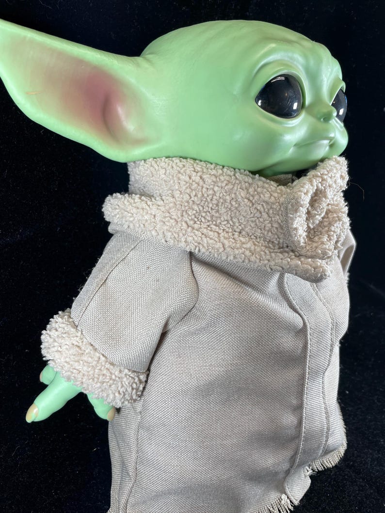 May include: A green-skinned doll with large black eyes and pointed ears. The doll is dressed in a tan coat with a fluffy, cream-coloured collar and cuffs. The doll's hand is extended, and the overall appearance is reminiscent of a popular character.