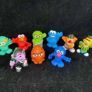 Puede incluir: Una colección de coloridas figuras de personajes de Barrio Sésamo. Las figuras incluyen a Elmo, Monstruo de las Galletas, Big Bird, Oscar el Gruñón y otros. Cada figura es de colores brillantes y de plástico.