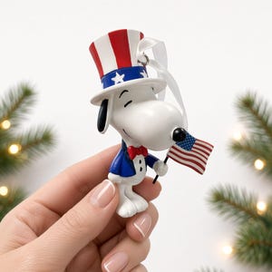 May include: A Snoopy figurine ornament wearing a red, white, and blue Uncle Sam top hat with stars, a blue jacket, and a red bow tie. The character holds a small American flag in one hand and features a white ribbon loop for hanging purposes.