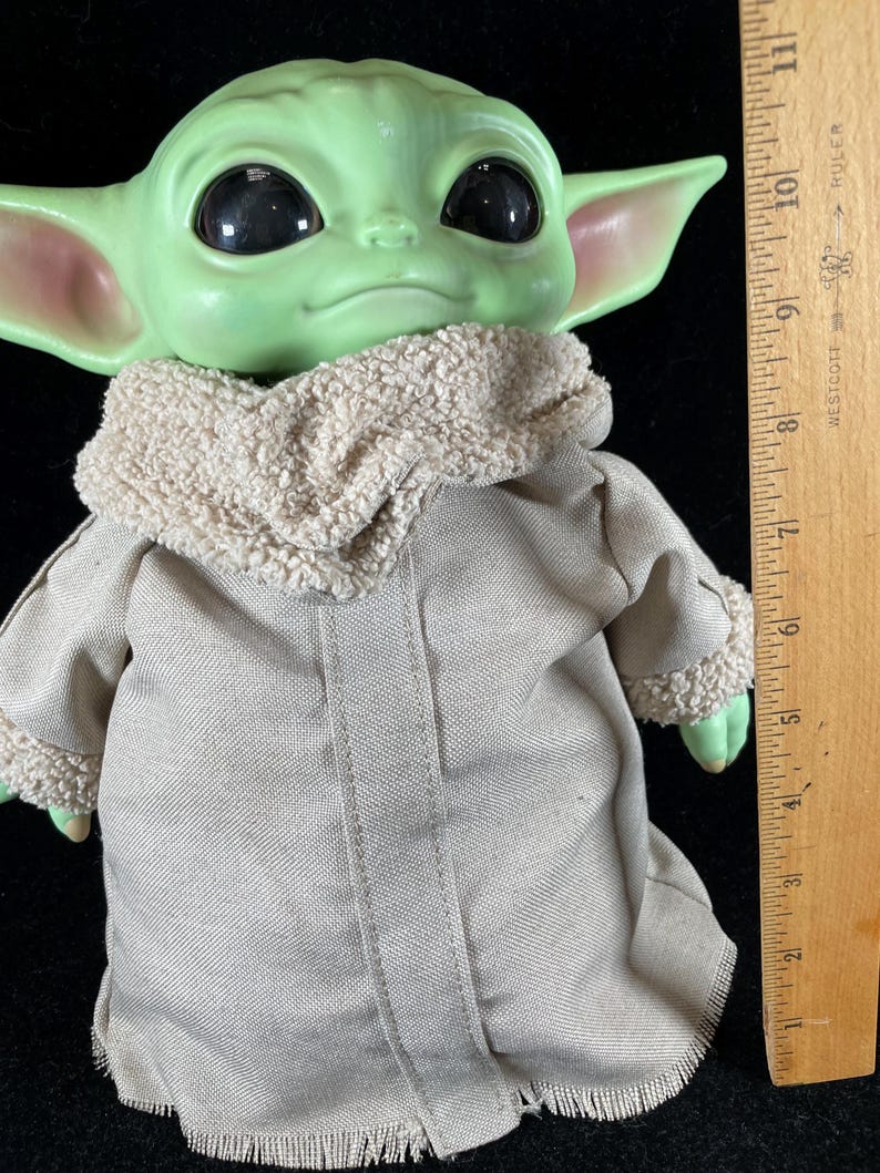 May include: A green-skinned doll with large black eyes and pointed ears. The doll is wearing a tan coat with a fuzzy collar. A wooden ruler is next to the doll, showing measurements in inches.