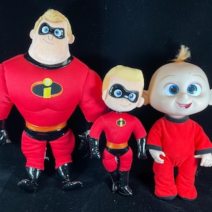 May include: Three plush toys from the Incredibles movie. The largest toy is Mr. Incredible, a red superhero with a black mask and the letter 'I' on his chest. The middle toy is Dash, a smaller red superhero with a black mask and the letter 'I' on his chest. The smallest toy is Jack-Jack, a baby in a red onesie.