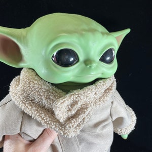 May include: A green plush toy with large eyes and a brown fur collar. The toy is wearing a tan jacket.