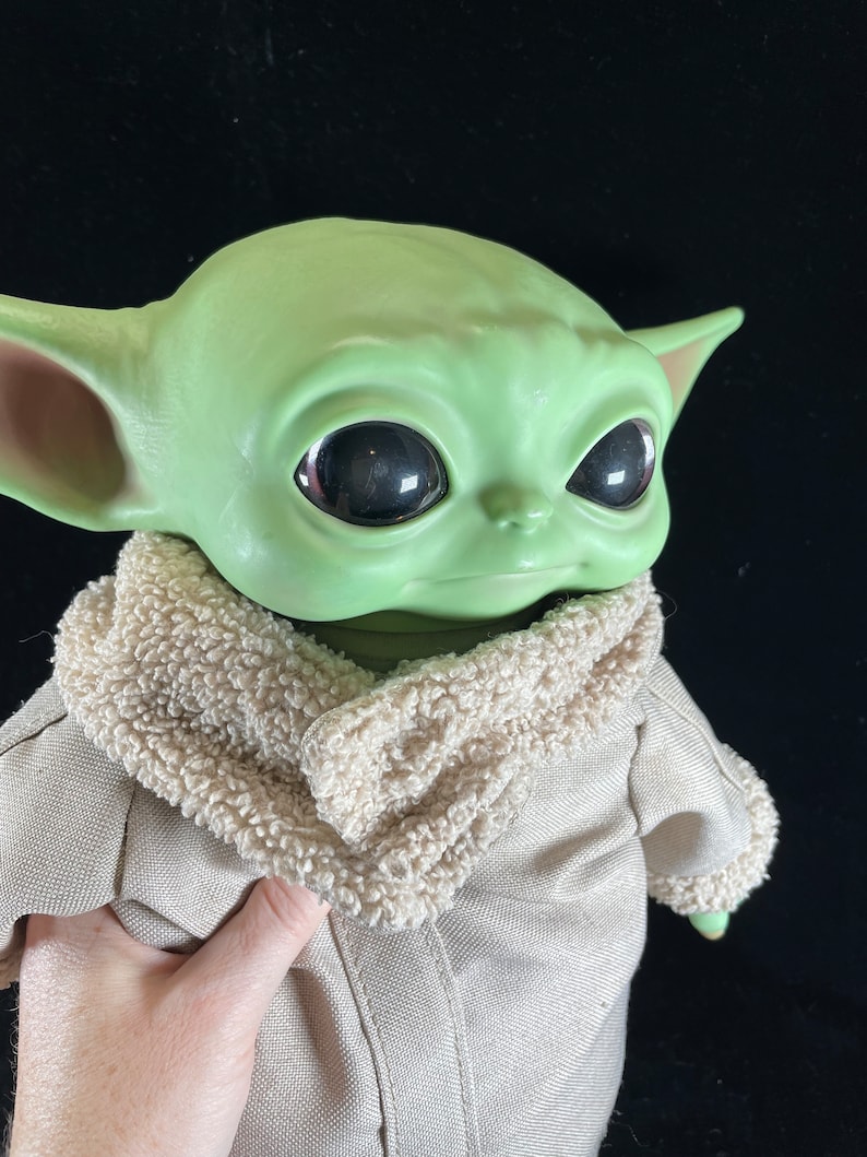 May include: A green plush toy with large eyes and a brown fur collar. The toy is wearing a tan jacket.