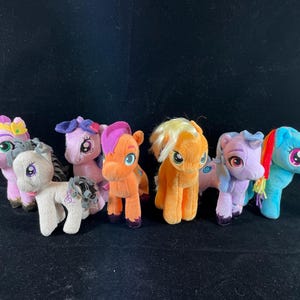 May include: Seven plush toy ponies in various colours, including purple, pink, orange, yellow, blue, and grey. The ponies have different coloured eyes and manes.