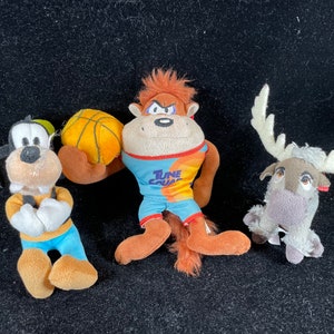 Puede incluir: Tres peluches: Goofy, un mono con una pelota de baloncesto y un reno. Goofy lleva una camisa amarilla, pantalones azules y un gorro verde. El mono lleva una camiseta con "Tune Squad". El reno tiene cuernos blancos.