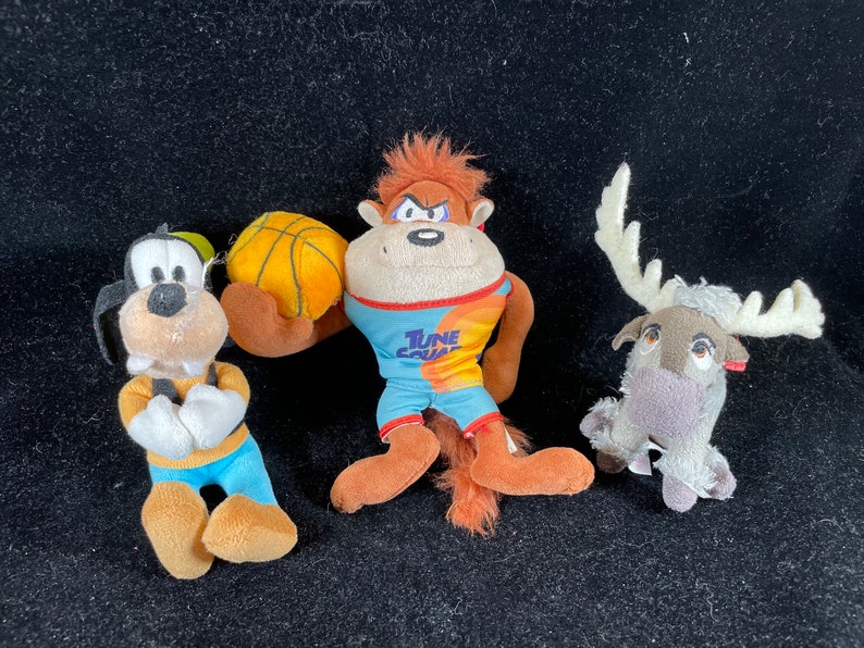 Puede incluir: Tres peluches: Goofy, un mono con una pelota de baloncesto y un reno. Goofy lleva una camisa amarilla, pantalones azules y un gorro verde. El mono lleva una camiseta con "Tune Squad". El reno tiene cuernos blancos.