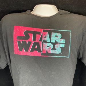 Op de afbeelding: Zwart T-shirt met het "STAR WARS" logo in een rechthoekig ontwerp. De bovenste helft van de tekst is rood en de onderste helft is turquoise. Het shirt wordt getoond op een witte mannequin.