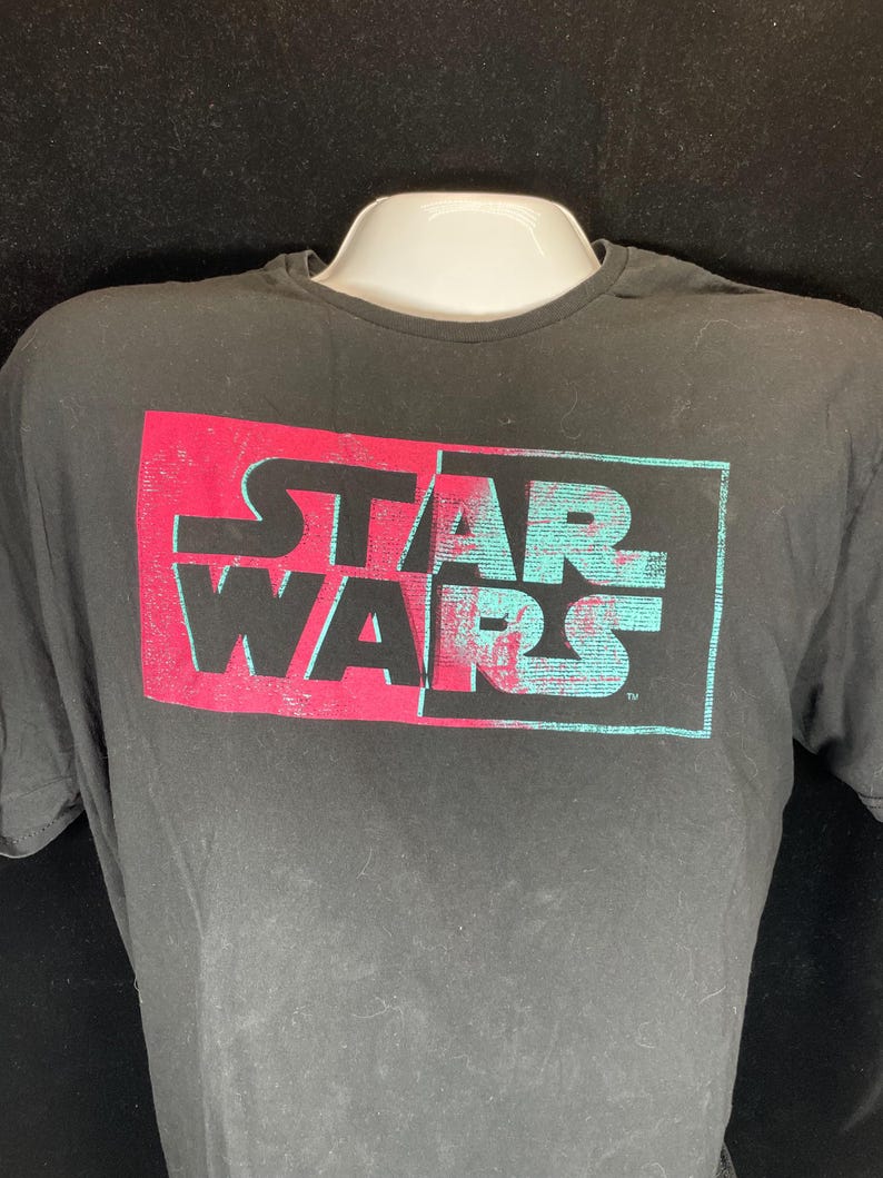 Op de afbeelding: Zwart T-shirt met het "STAR WARS" logo in een rechthoekig ontwerp. De bovenste helft van de tekst is rood en de onderste helft is turquoise. Het shirt wordt getoond op een witte mannequin.