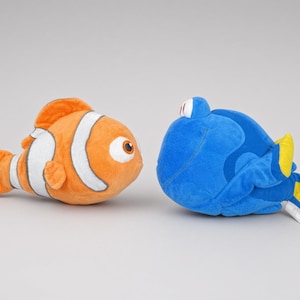 May include: Two plush fish toys facing each other. One is an orange clownfish with white vertical stripes and large eyes. The other is a blue tang fish with yellow fins and tail, featuring a rounded body shape and large white eyes with black pupils.