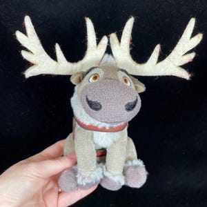 May include: A plush reindeer toy with large, cream-colored antlers with red accents. The reindeer has a brown nose, black eyes, and a red and brown harness. The toy is tan and gray with fluffy accents on the legs and chest.