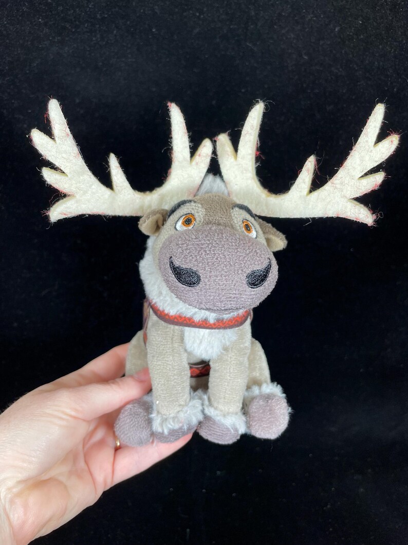 May include: A plush reindeer toy with large, cream-colored antlers with red accents. The reindeer has a brown nose, black eyes, and a red and brown harness. The toy is tan and gray with fluffy accents on the legs and chest.