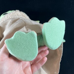 May include: A plush toy of a green character with a brown robe and large green feet. The toy is being held by a hand.