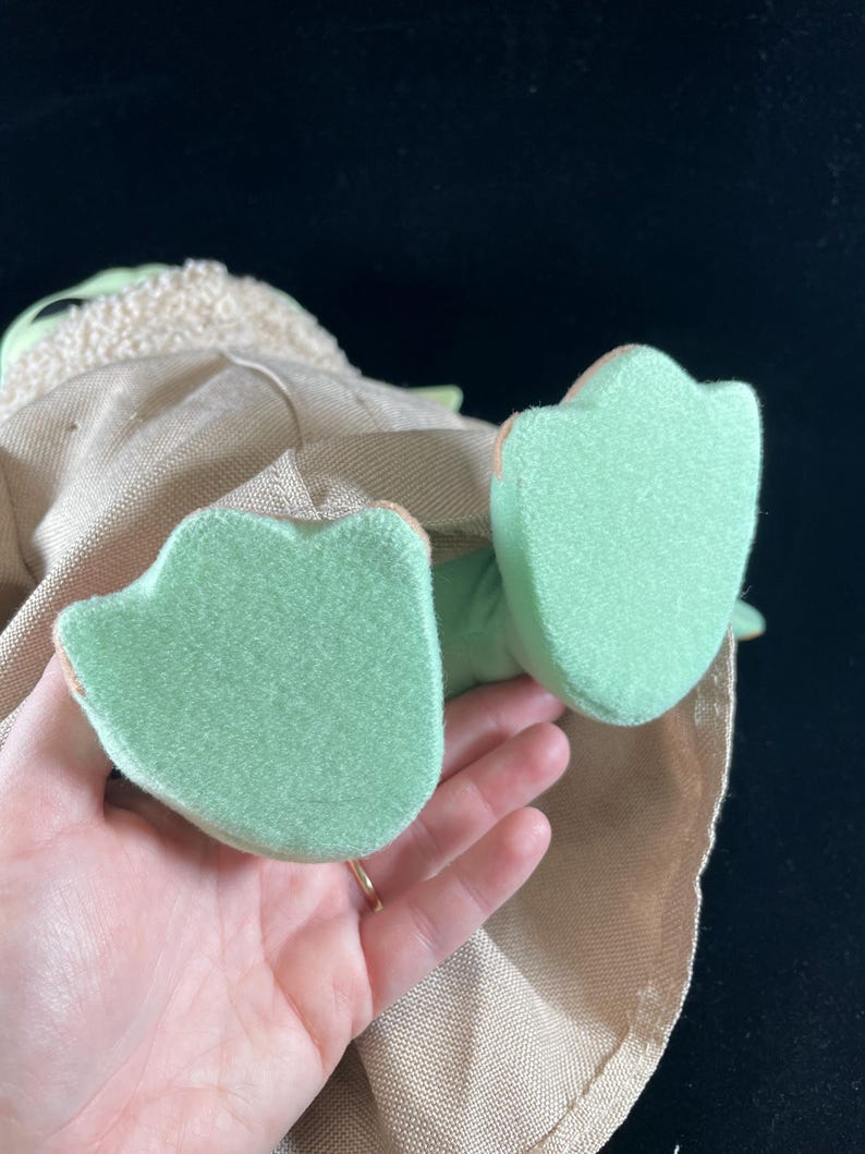 May include: A plush toy of a green character with a brown robe and large green feet. The toy is being held by a hand.