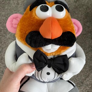 May include: A plush Mr. Potato Head toy wearing a gray hat, a black bow tie, and a gray vest over a white shirt. The toy has large white eyes, an orange nose, pink ears, and a black mustache.