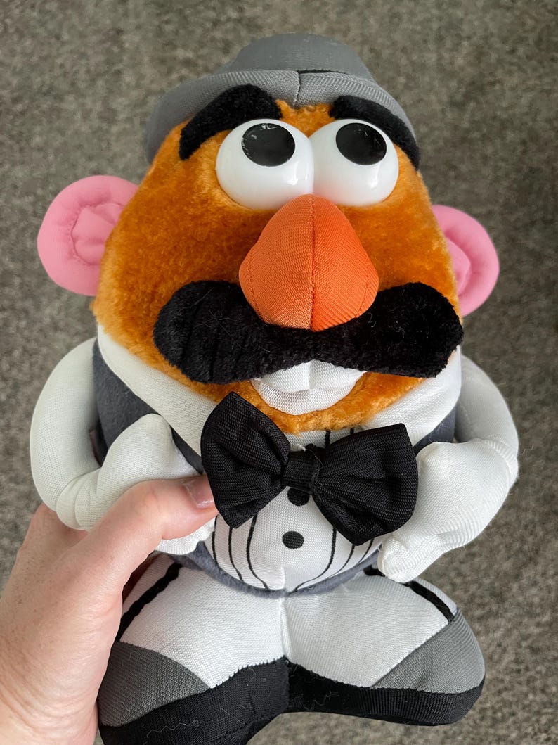 May include: A plush Mr. Potato Head toy wearing a gray hat, a black bow tie, and a gray vest over a white shirt. The toy has large white eyes, an orange nose, pink ears, and a black mustache.