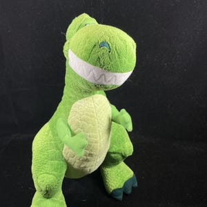 May include: A plush green Tyrannosaurus Rex toy with a friendly expression. The dinosaur has a light green belly, dark green feet, and a wide, white-toothed grin. The toy is made of soft, textured fabric.