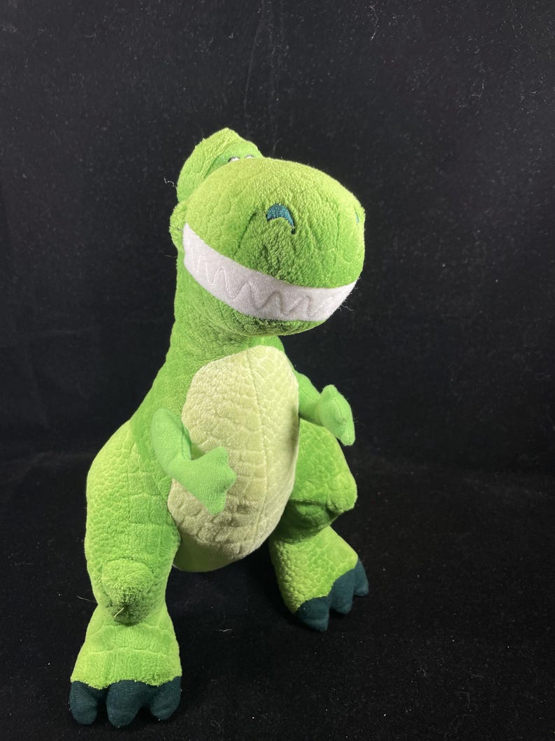 May include: A plush green Tyrannosaurus Rex toy with a friendly expression. The dinosaur has a light green belly, dark green feet, and a wide, white-toothed grin. The toy is made of soft, textured fabric.