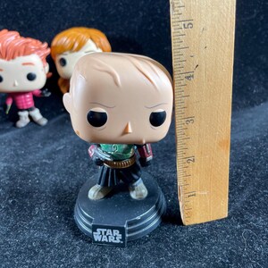 May include: A Star Wars Funko Pop figure of a character in a green and black outfit, standing on a black base with the Star Wars logo. A wooden ruler is next to the figure, measuring approximately 15 cm.