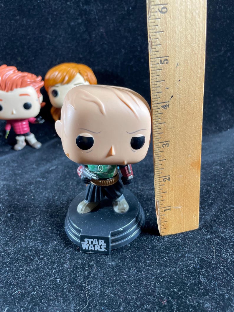 May include: A Star Wars Funko Pop figure of a character in a green and black outfit, standing on a black base with the Star Wars logo. A wooden ruler is next to the figure, measuring approximately 15 cm.