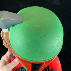May include: A plush toy with a green helmet and red armour, resembling a science fiction character. The toy has a grey and gold accessory attached to the helmet. The toy is held against a black background.