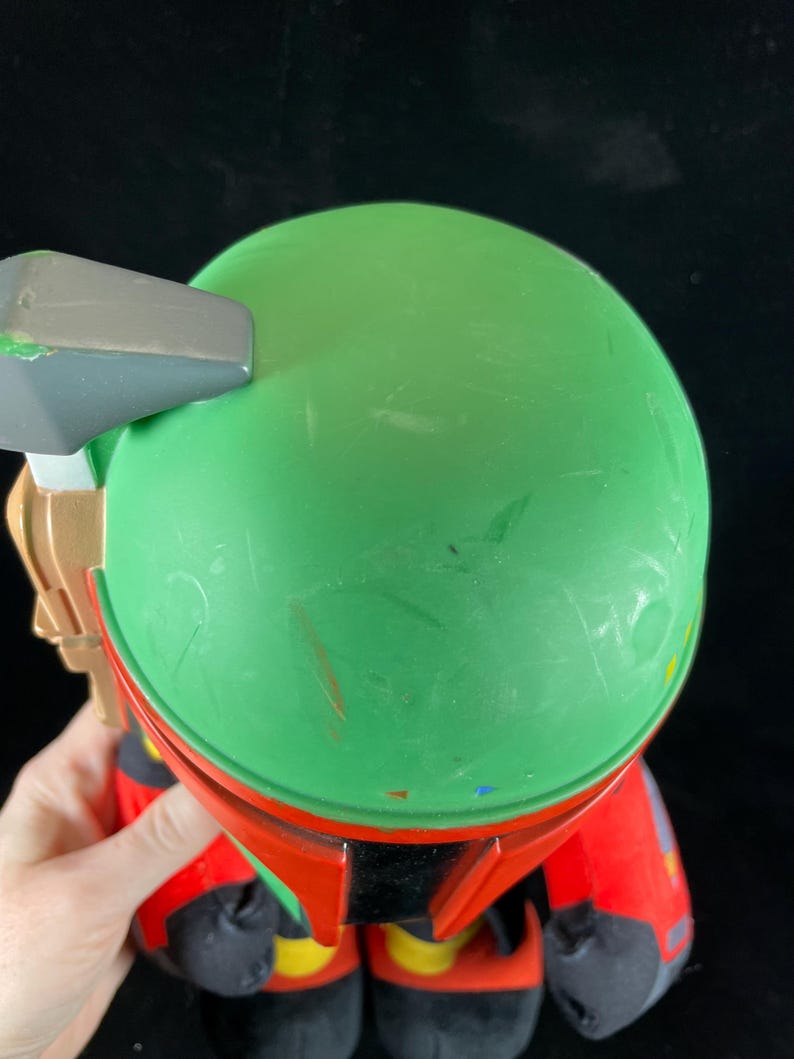 May include: A plush toy with a green helmet and red armour, resembling a science fiction character. The toy has a grey and gold accessory attached to the helmet. The toy is held against a black background.
