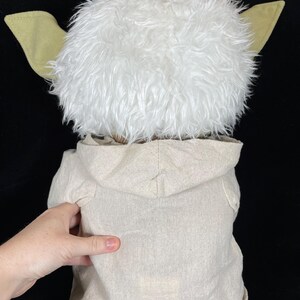 Op de afbeelding: Een Yoda-knuffel, van achteren gezien, met een pluizige witte kop, grote groene oren en een beige capuchon. De voeten van de pop zijn ook groen. De achtergrond is zwart.