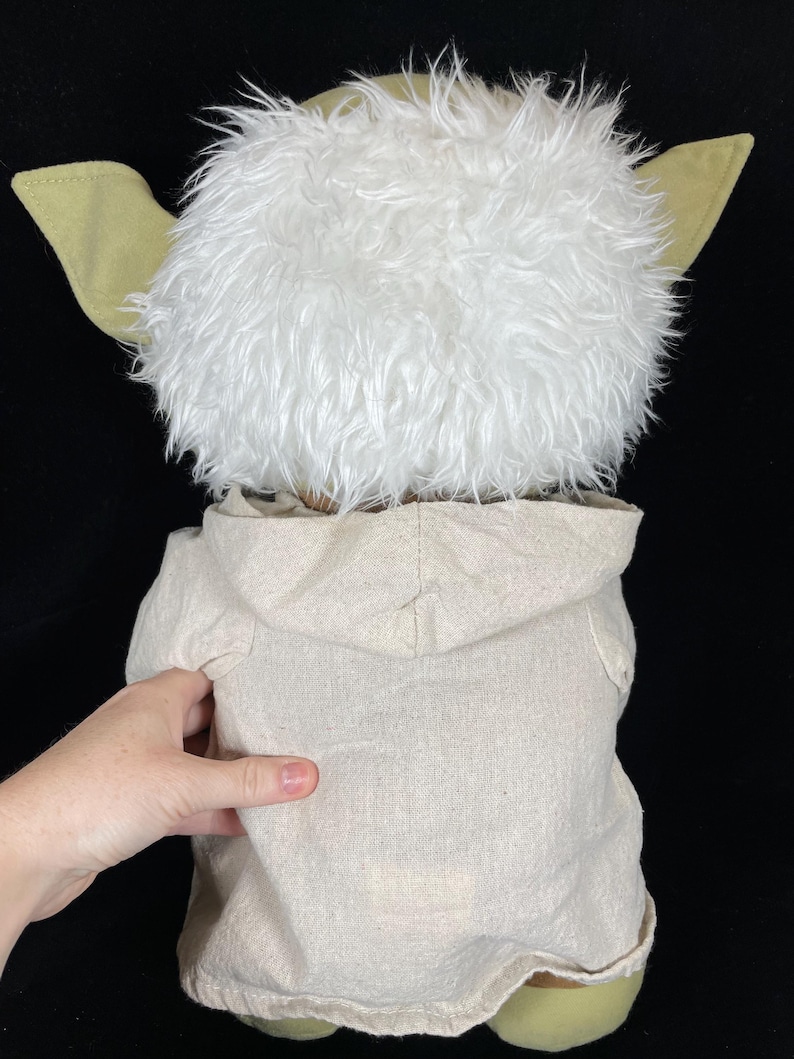 Op de afbeelding: Een Yoda-knuffel, van achteren gezien, met een pluizige witte kop, grote groene oren en een beige capuchon. De voeten van de pop zijn ook groen. De achtergrond is zwart.