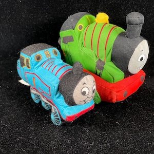 Puede incluir: Dos trenes de juguete de peluche, uno azul y otro verde, están uno al lado del otro. El tren azul tiene rayas rojas y el número 1. El tren verde tiene rayas rojas y una chimenea negra. Ambos tienen caras sonrientes.