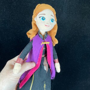 May include: A plush doll of a character with orange hair, blue eyes, and a smiling expression. The doll wears a black dress with gold accents, a purple cape, and a pink trim. The doll is held against a black background.