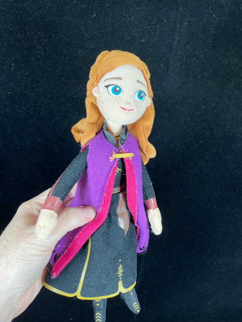 May include: A plush doll of a character with orange hair, blue eyes, and a smiling expression. The doll wears a black dress with gold accents, a purple cape, and a pink trim. The doll is held against a black background.
