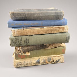 Puede incluir: Una pila vertical de siete libros de tapa dura antiguos con bordes desgastados y deshilachados, con cubiertas de tela descolorida en tonos gris, azul, beige y verde oliva. Los lomos muestran varios títulos y nombres de autores, incluido The Case of Richard Meynell.
