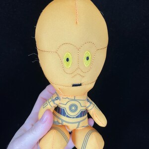 Op de afbeelding: Een pluche speelgoed van het Star Wars-personage C-3PO. De pop is voornamelijk geel met zwarte en grijze details. Het heeft geborduurde kenmerken, waaronder gele ogen en een zwarte mond. Het speelgoed wordt door een hand vastgehouden.